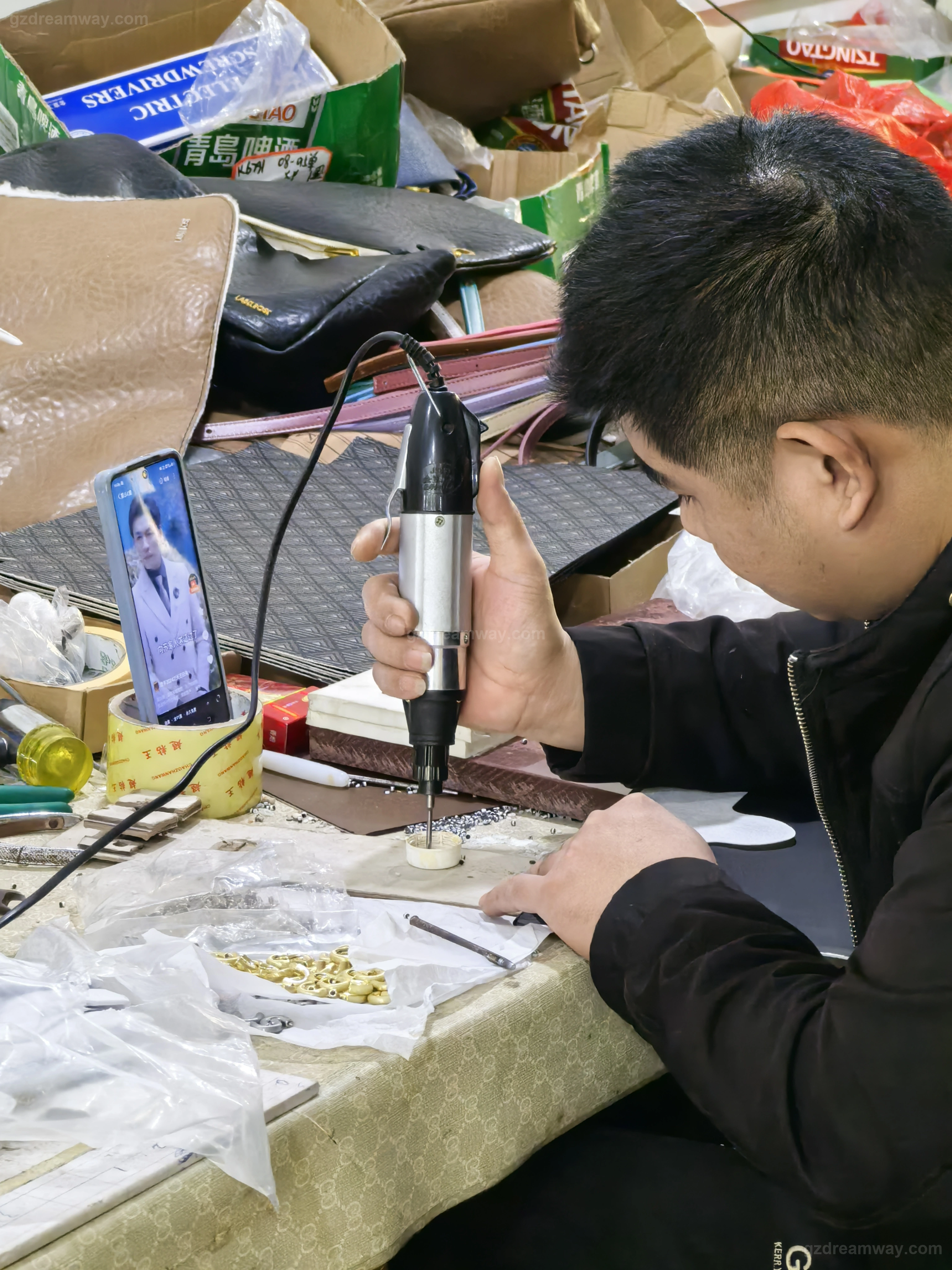 Worker using an electric screwdriver to install brass bottom studs, metal buckles and magnetic closures on a custom leather tote bag at hardware assembly station for private-label bag OEM production