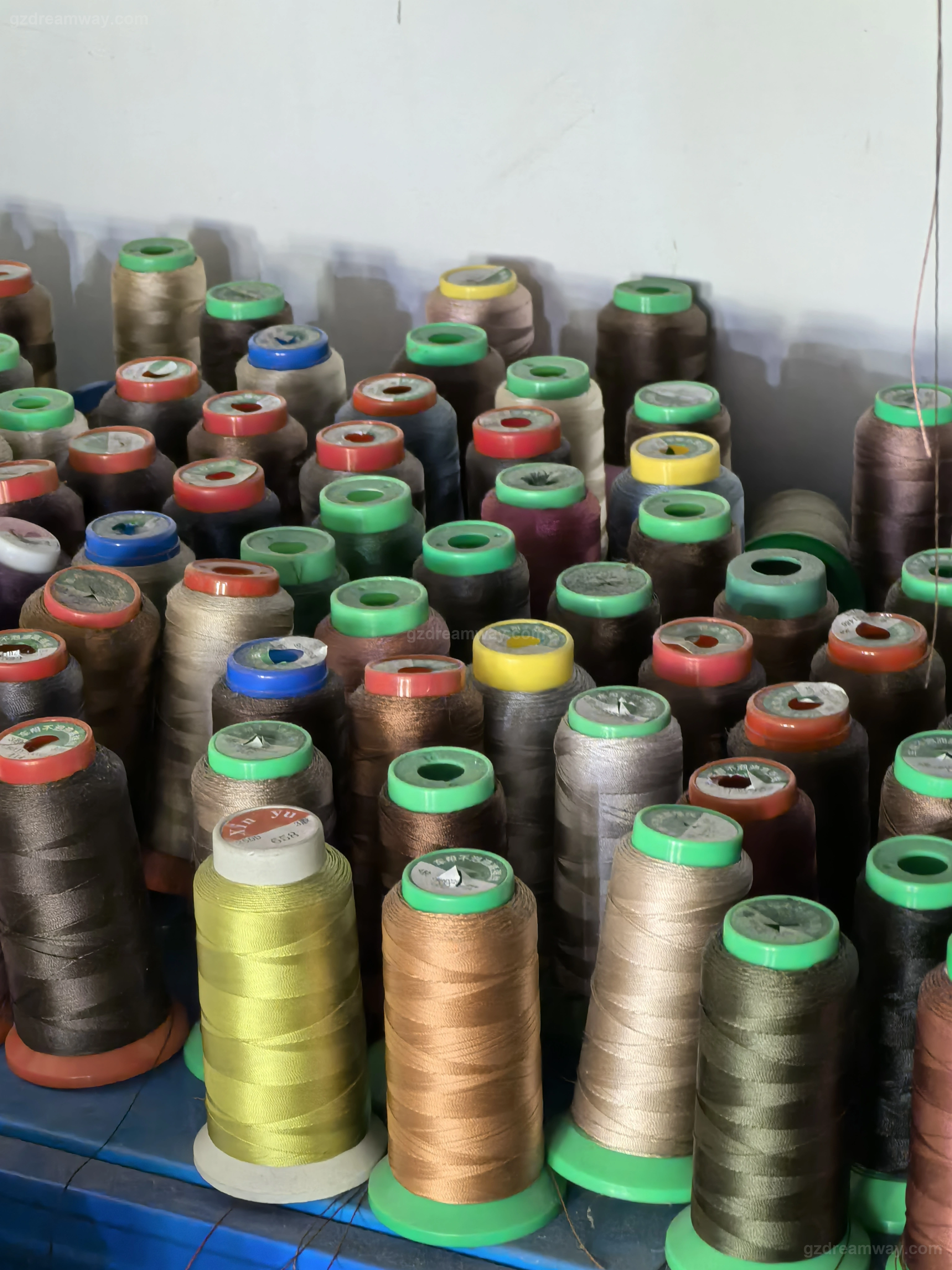 Color-matched bonded nylon and polyester sewing thread spools organized on a wall rack next to leather bag, canvas tote bag and custom backpack sewing machines at GZ Dreamway production line