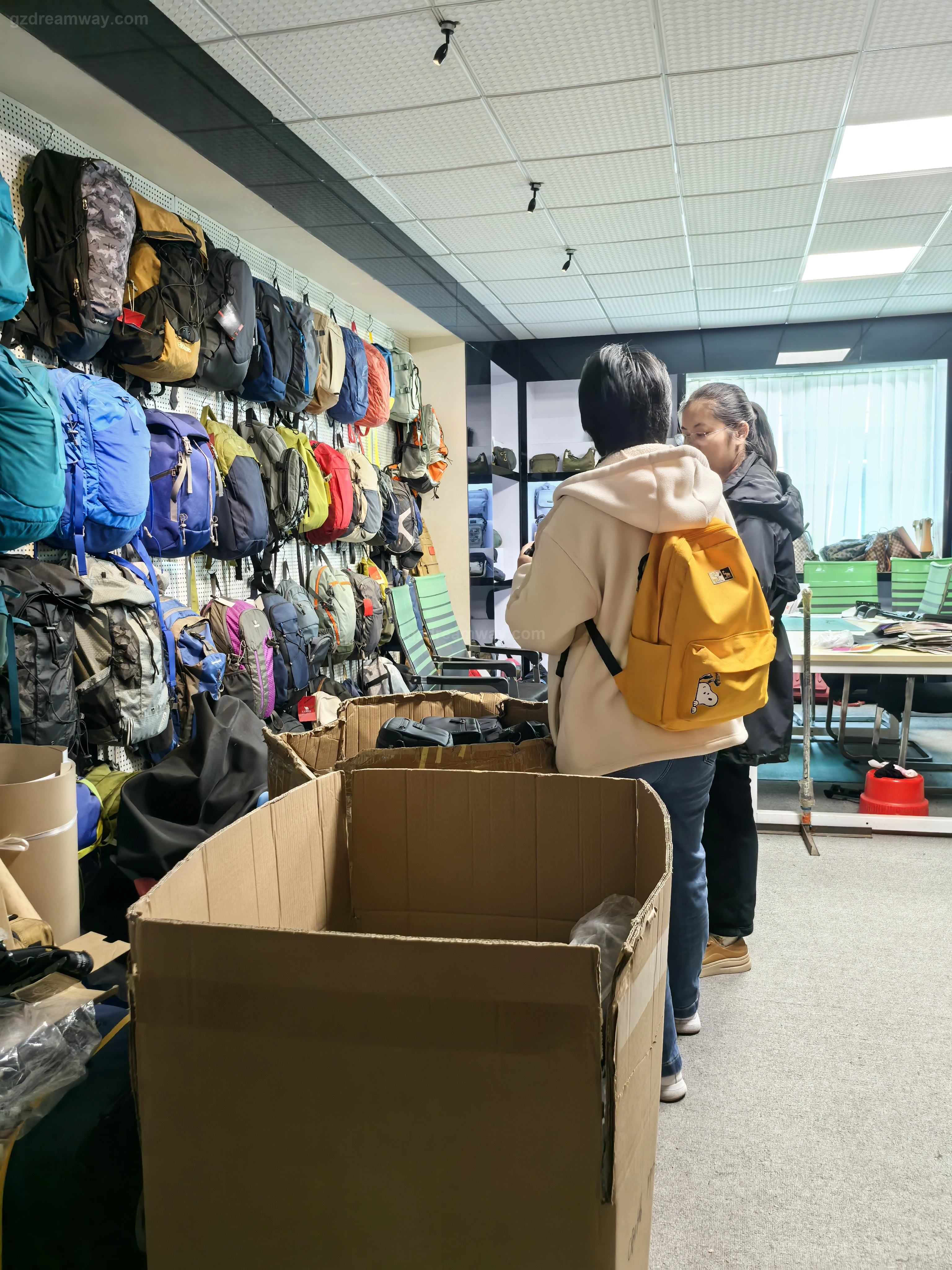 Factory showroom where overseas clients fit a yellow canvas school bag backpack beside a wall of outdoor travel bag, custom leather bag and tote bag prototypes alongside pattern development engineers at GZ Dreamway