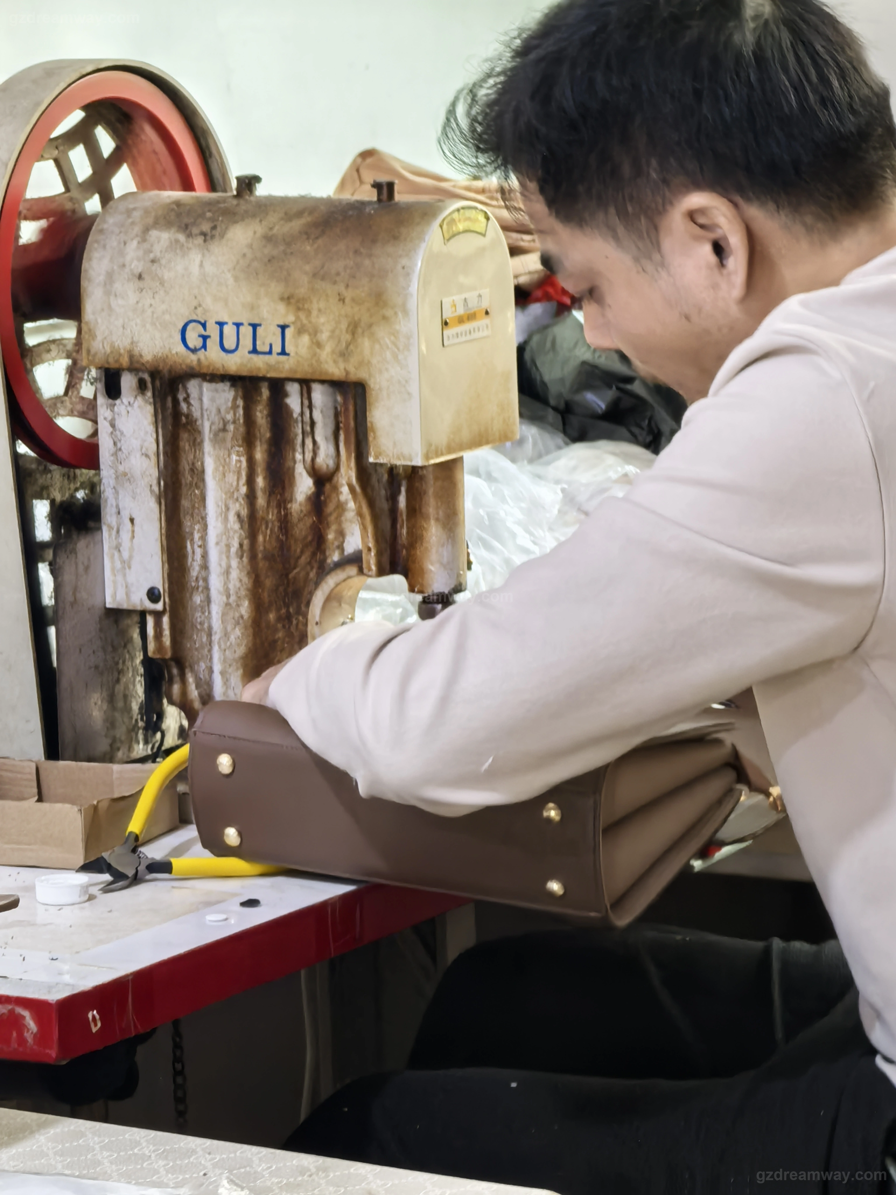 Worker using a pneumatic riveting machine to install protective metal bottom studs on a dark brown leather handbag tote at GZ Dreamway Guangzhou factory hardware assembly station